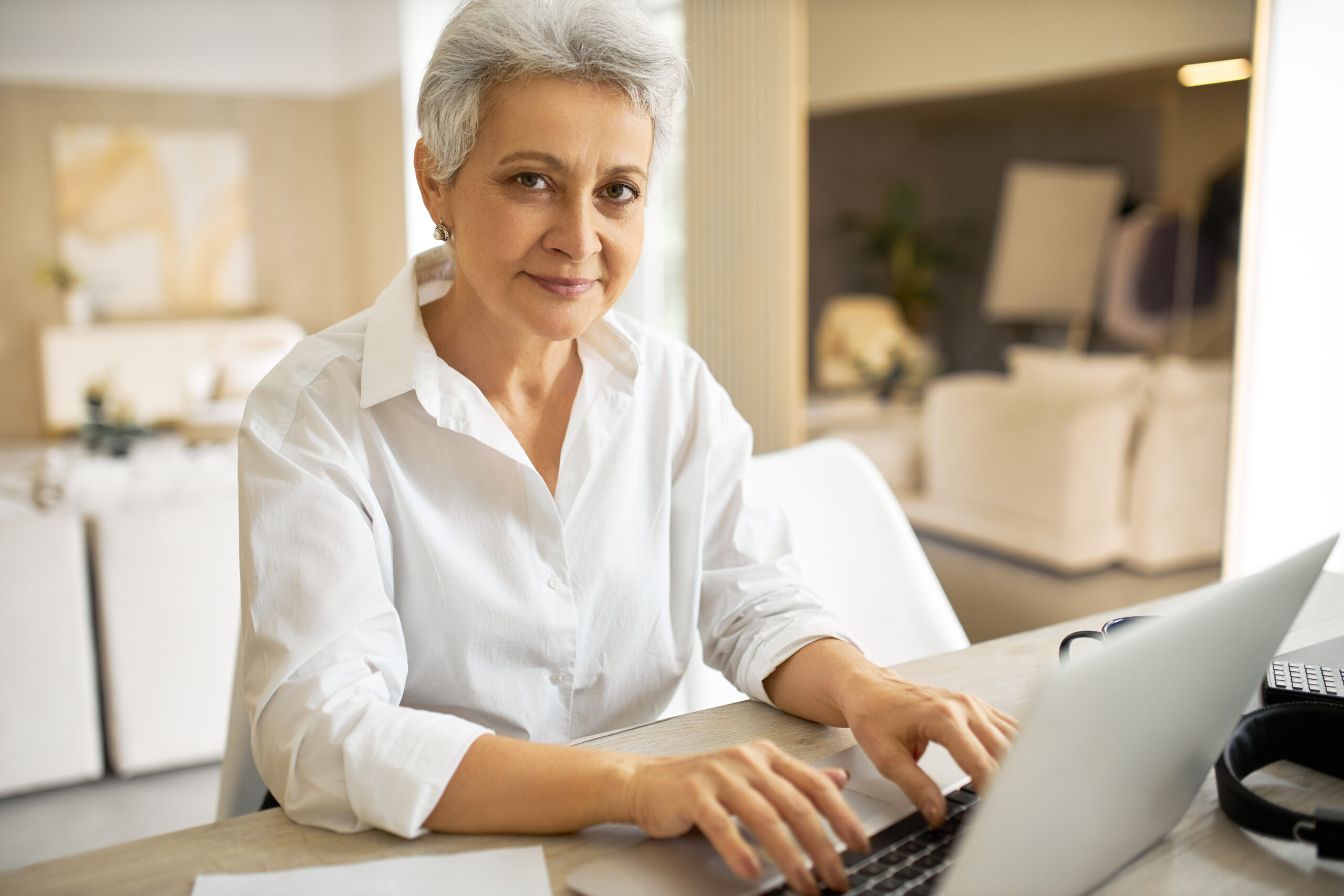 Portrait of fashionable cheerful 50 year old woman writer in white shirt using generic electronic gadget for work, typing another chapter of her new book, having happy look full of inspiration